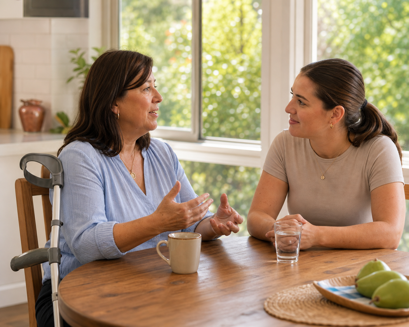 A participant using a forearm crutch speaks while a support worker listens at a kitchen table.